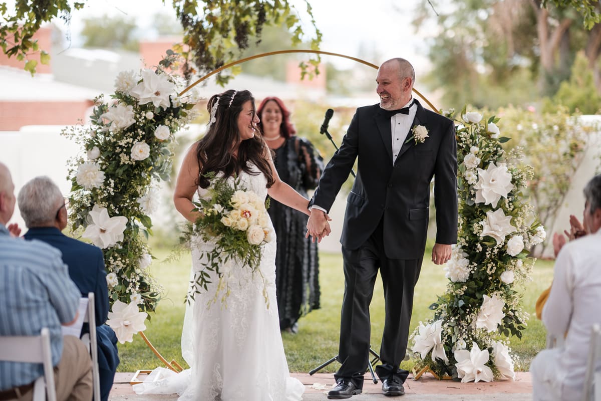 Claudia and Carlos walk back up the aisle under a floral arch at El Verde Inn, Albuquerque