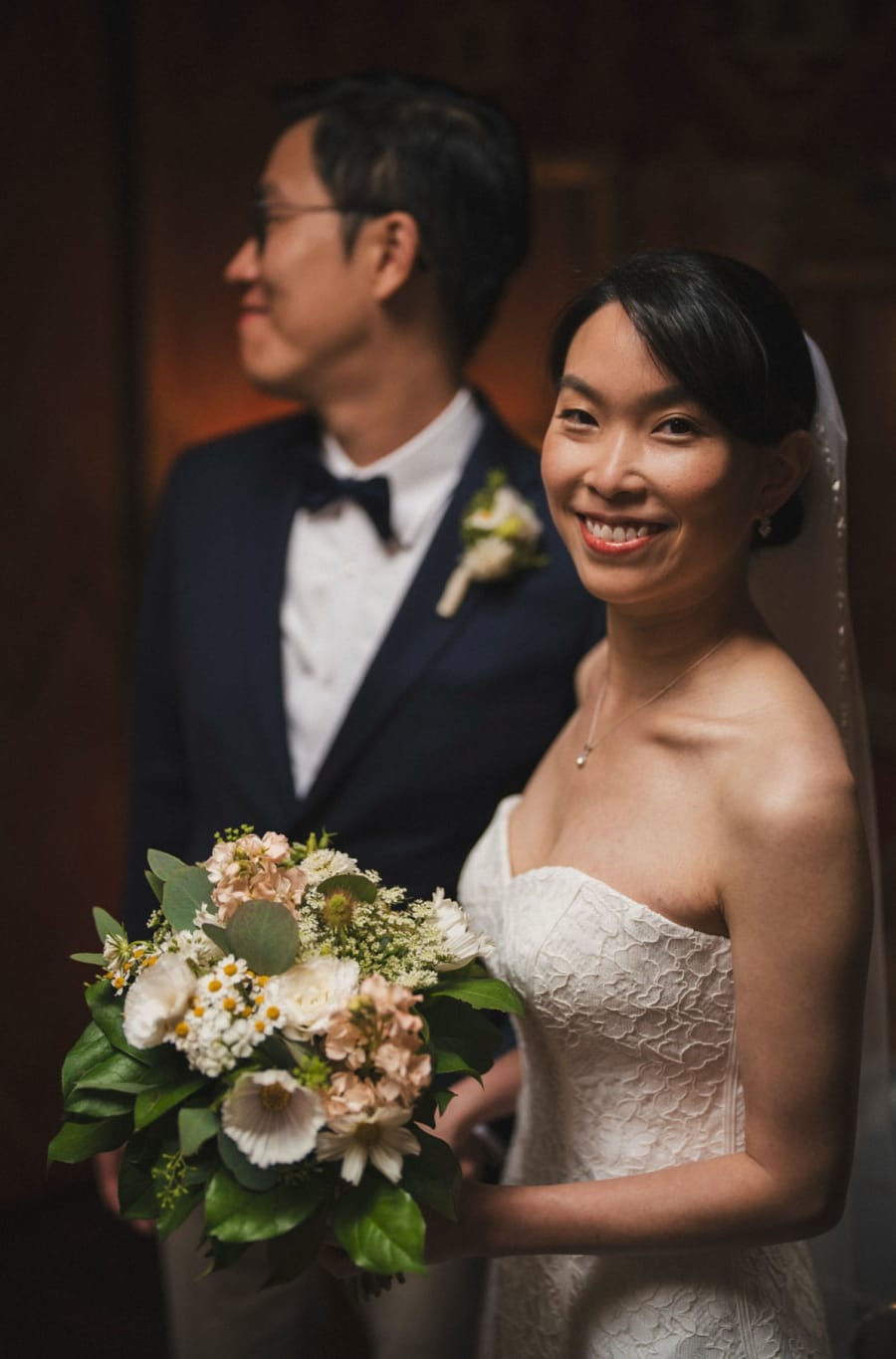 Bride smiling over her bouquet, groom behind her in soft focus