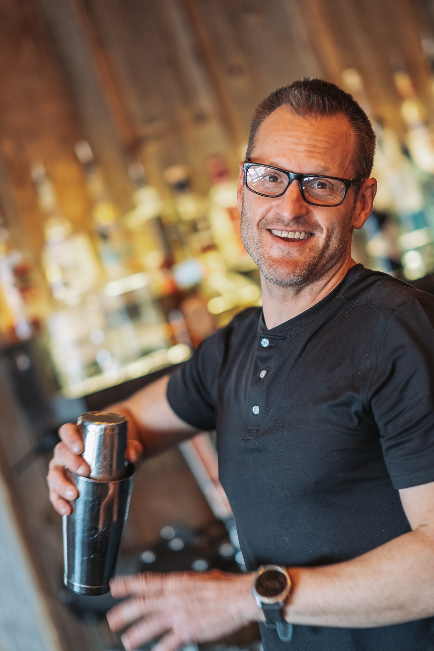 This portrait documents a bartender or mixologist in a contemporary indoor setting, holding a stainless steel cocktail shaker with a warm, confident smile — Casey Addason Photography