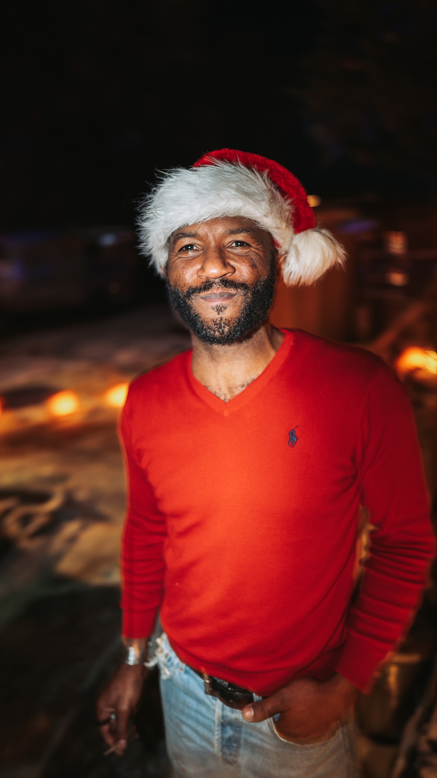 This casual outdoor portrait documents a man in festive attire standing on a beach at night, wearing a red long-sleeve polo shirt, light-wash jeans, and a red Santa hat with white fur trim — Casey Addason Photography