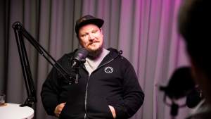 Mustaan huppariin ja lippikseen pukeutunut valkohattuhakkeri Benjamin Särkkä puhuu podcast-mikrofoniin studiossa, jossa on violetti valaistus.