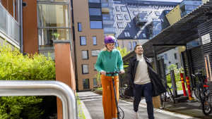 Skootterilla ajava henkilö, jolla on kypärä päässään, kun toinen henkilö kävelee hänen vieressään modernilla kaupunkialueella.