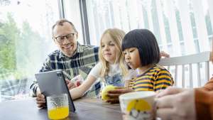 Mies ja kaksi lasta istuvat pöydän ääressä, katsovat yhdessä tablettia, ja heidän edessään on juomia.