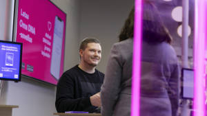 Mies hymyilee auttaessaan naista teknisen kaupan tiskillä, jonka taustalla on kirkkaan vaaleanpunainen kyltti.