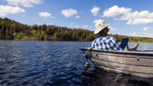 Ruudulliseen paitaan pukeutunut henkilö työskentelee kannettavalla tietokoneella istuessaan veneessä järvellä puiden ympäröimänä.