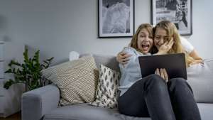 Kaksi naista sohvalla nauramassa ja halailemassa, kun he katsovat yhdessä kannettavan tietokoneen näyttöä.