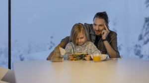 Mies ja lapsi istuvat pöydän ääressä; lapsi pelaa laitteella, kun mies puhuu puhelimeen.