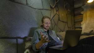 Mies istuu sohvalla, hymyilee ja elehtii käyttäessään kannettavaa tietokonetta hämärässä huoneessa, jossa on kiviseinät.