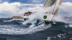 Purjevene leikkaa kovia aaltoja, miehistö nojaa laidan yli; taustalla myrskyinen taivas ja kallioinen maa.