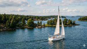 Valkoinen purjevene liukuu tyynessä sinisessä vedessä metsäisen saaren lähellä osittain pilvisenä päivänä.