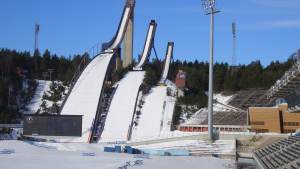 Kolme suurta hyppyrimäkeä lumisella mäellä, katsomot ja valonheittimet kirkkaan sinisen taivaan alla.