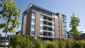 Moderni kerrostalo, jossa on parvekkeet, suuret ikkunat ja puita etualalla aurinkoisena päivänä.