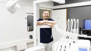 Hymyilevä hammaslääkäri seisoo modernissa hammashoitolassa, jossa on laitteita ja tietokone näkyvissä.