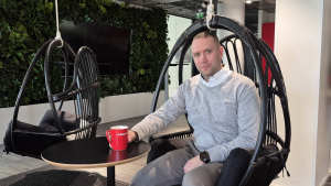 Mies istuu riipputuolilla modernissa toimistossa ja pitelee punaista mukia pienen pyöreän pöydän ääressä.