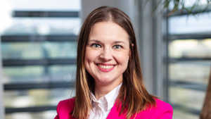 Kirkkaan vaaleanpunaiseen bleiseriin pukeutunut nainen hymyilee, kun hän seisoo sisätiloissa ja taustalla on sumeat ikkunat.