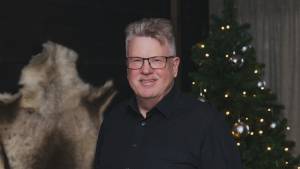 Hymyilevä mustapaitainen mies seisoo koristellun joulukuusen ja seinällä olevan turkismaton lähellä.