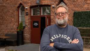 Silmälasipäinen ja parrakas mies seisoo kädet ristissä tiilirakennuksen ulkopuolella, jonka kyltissä lukee Wool Factory Bistro.