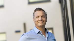 Hymyilevä vaaleansiniseen paitaan pukeutunut mies seisoo ulkona modernin rakennuksen edessä ja katsoo kameraan.