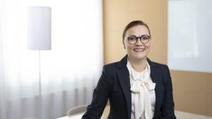 Puvussa ja silmälaseissa oleva nainen hymyilee valoisassa, modernissa toimistoympäristössä, jossa on lamppu ja valkotaulu.