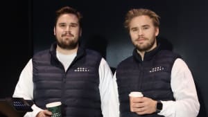 Kaksi miestä, joilla on samanlaiset liivit ja kahvikupit kädessään, seisovat sisätiloissa kameraan päin.