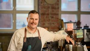 Esiliinaan pukeutunut mies hymyilee kahvimyllyjen vieressä viihtyisässä kahvilassa, jossa on tiiliseinät ja suuret ikkunat.