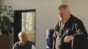 Kaksi miestä toimistossa, toinen istuu ja hymyilee toiselle, joka seisoo ja puhuu, molemmat tietokoneiden ääressä.