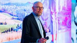 Kalju, silmälasipäinen mies puhuu lavalla kaukosäädin kädessään, ja hänen takanaan on värikäs tausta.