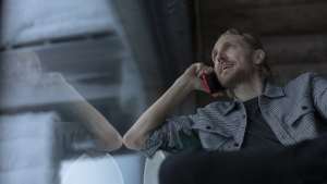 Parrakas mies puhuu punaiseen puhelimeen istuessaan sisätiloissa heijastavan lasi-ikkunan äärellä.