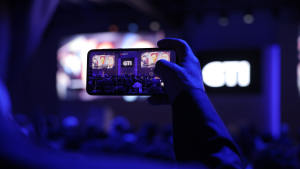Henkilö nauhoittaa lavaesitystä puhelimellaan täpötäydessä, sinisellä valaistussa tilaisuudessa.