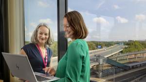 Kaksi naista seisoo ikkunan ääressä hymyillen ja katselee kannettavaa tietokonetta, jonka ulkopuolella on valtatie ja sininen taivas.