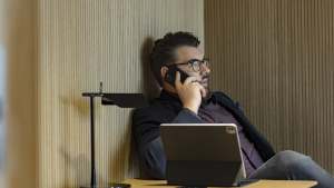 Silmälasipäinen mies istuu työpöydän ääressä, puhuu puhelimeen, edessä on tabletti ja lamppu.