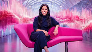 Tummiin vaatteisiin pukeutunut nainen istuu modernilla vaaleanpunaisella sohvalla, jonka taustalla on elinvoimainen vaaleanpunainen abstrakti taide.