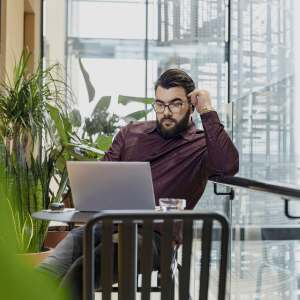 Silmälasipäinen mies työskentelee kannettavalla tietokoneella modernissa sisätyötilassa, jossa on kasveja ja suuria ikkunoita.