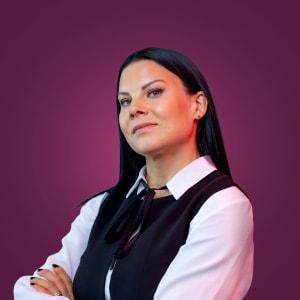 Nainen, jolla on pitkät tummat hiukset, musta liivi ja valkoinen paita, seisoo itsevarmasti violettia taustaa vasten.