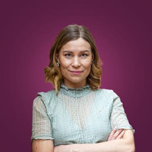 Nainen, jolla on hartioiden pituiset vaaleat hiukset ja vaaleanvihreä pusero, seisoo kädet ristissä violetilla taustalla.