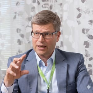 Siniseen pukuun ja silmälaseihin pukeutunut mies, joka istuu sisätiloissa valkoisilla verhoilla verhotun ikkunan lähellä, tekee eleitä puhuessaan.