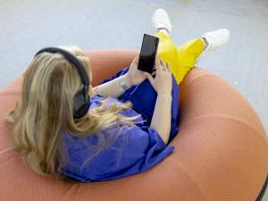 Henkilö, jolla on kuulokkeet ja joka rentoutuu oranssilla papusäkillä, katselee älypuhelinta, jalat ristissä.