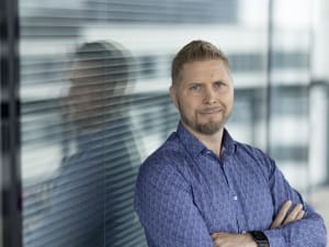 Sinipaitainen mies seisoo kädet ristissä nojaten lasiseinään, jossa näkyy heijastus.