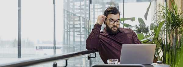 Silmälasipäinen mies työskentelee kannettavalla tietokoneella modernissa sisätyötilassa, jossa on kasveja ja suuria ikkunoita.