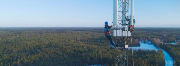 Kaksi turvavarusteisiin pukeutunutta työntekijää kiipeää korkeaa tukiasemaa lumisen metsämaiseman yllä kirkkaan sinisen taivaan alla.