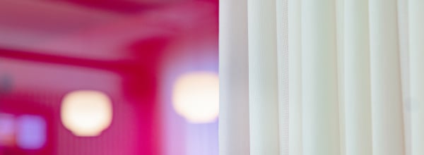 Valkoinen verho tarkennettuna oikealla, taustalla sumea vaaleanpunainen huone ja valot.
