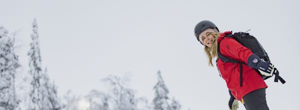 Hymyilevä nainen punaisessa takissa ja kypärässä lumilautailee lumisessa metsäisessä maisemassa.