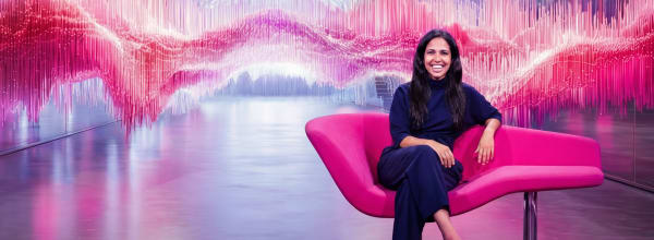 Tummiin vaatteisiin pukeutunut nainen istuu vaaleanpunaisella tuolilla, jonka taustalla on värikkäitä vaaleanpunaisia ja violetteja abstrakteja aaltoja.