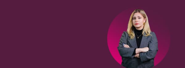 Vaaleatukkainen nainen harmaassa bleiserissä seisoo kädet ristissä violetilla taustalla.