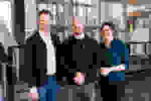 Kolme ihmistä seisoo ja hymyilee modernissa toimiston aulassa, jossa on lasiseinät ja portaikko.