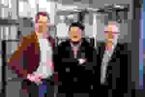 Kolme miestä seisoo yhdessä sisätiloissa hymyillen kameralle modernissa, lasiseinäisessä rakennuksessa.