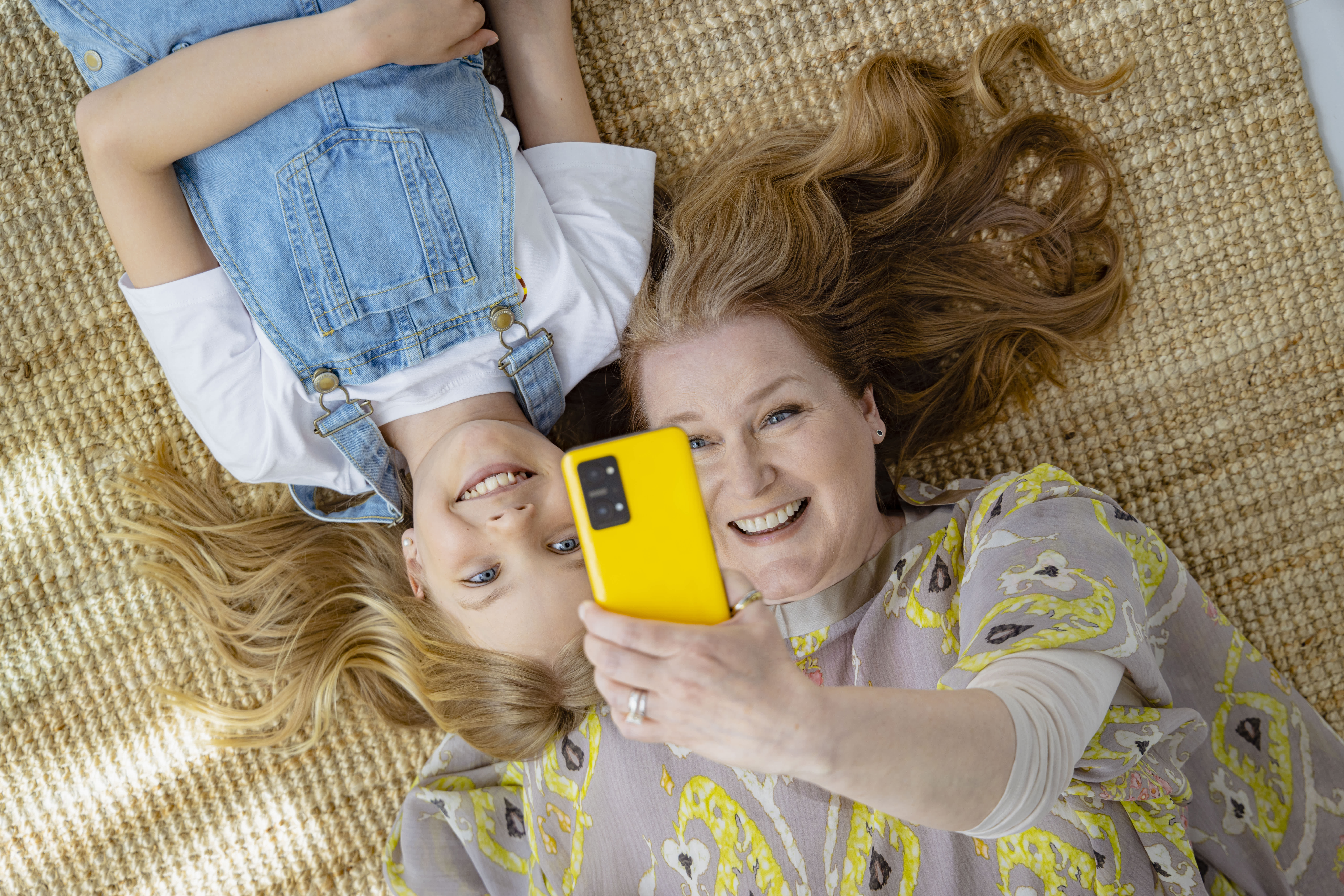 Nainen ja nuori tyttö makaavat matolla, hymyilevät ja ottavat selfien yhdessä.
