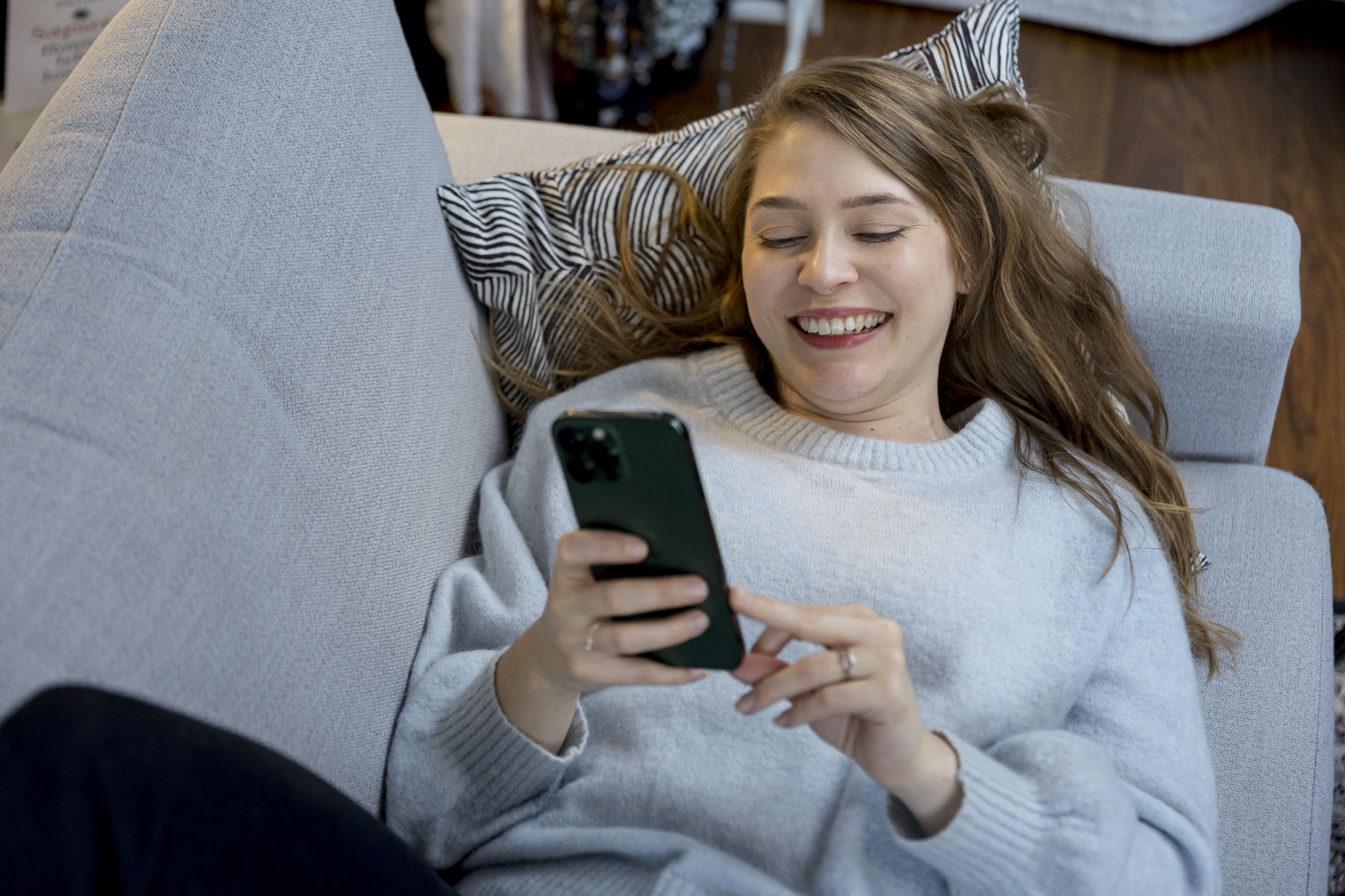 Nainen makaa sohvalla, hymyilee ja katselee älypuhelinta yllään vaaleanharmaa villapaita.