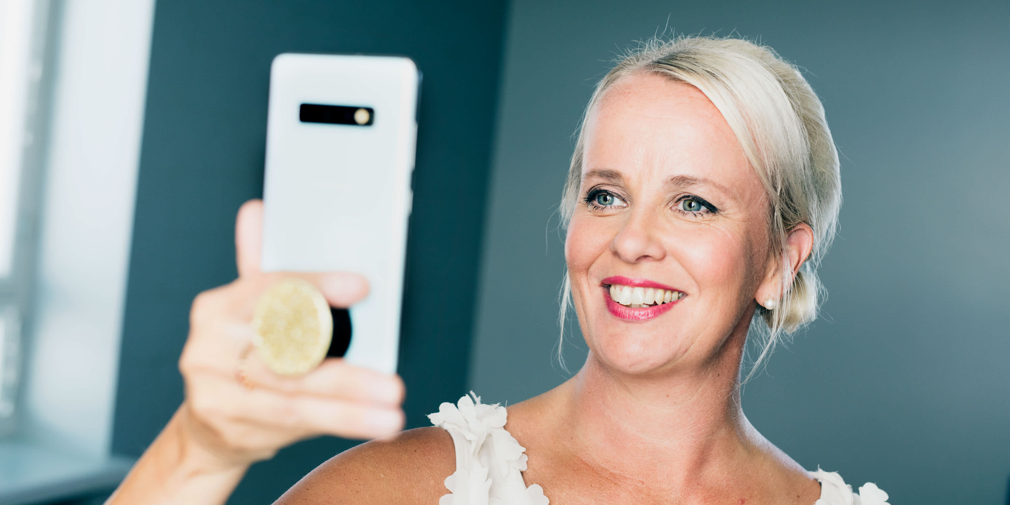 Hymyilevä vaaleatukkainen nainen ottaa selfien älypuhelimellaan kirkkaassa huoneessa.