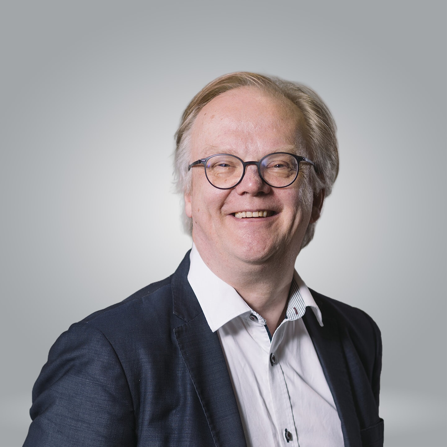 Hymyilevä silmälasipäinen mies, jolla on puvuntakki ja valkoinen paita harmaata taustaa vasten.